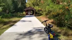 Stonecrest, GA image Paved trail leading to a wooden covered bridge surrounded by trees, with a bicycle and bags on the right side of the path
